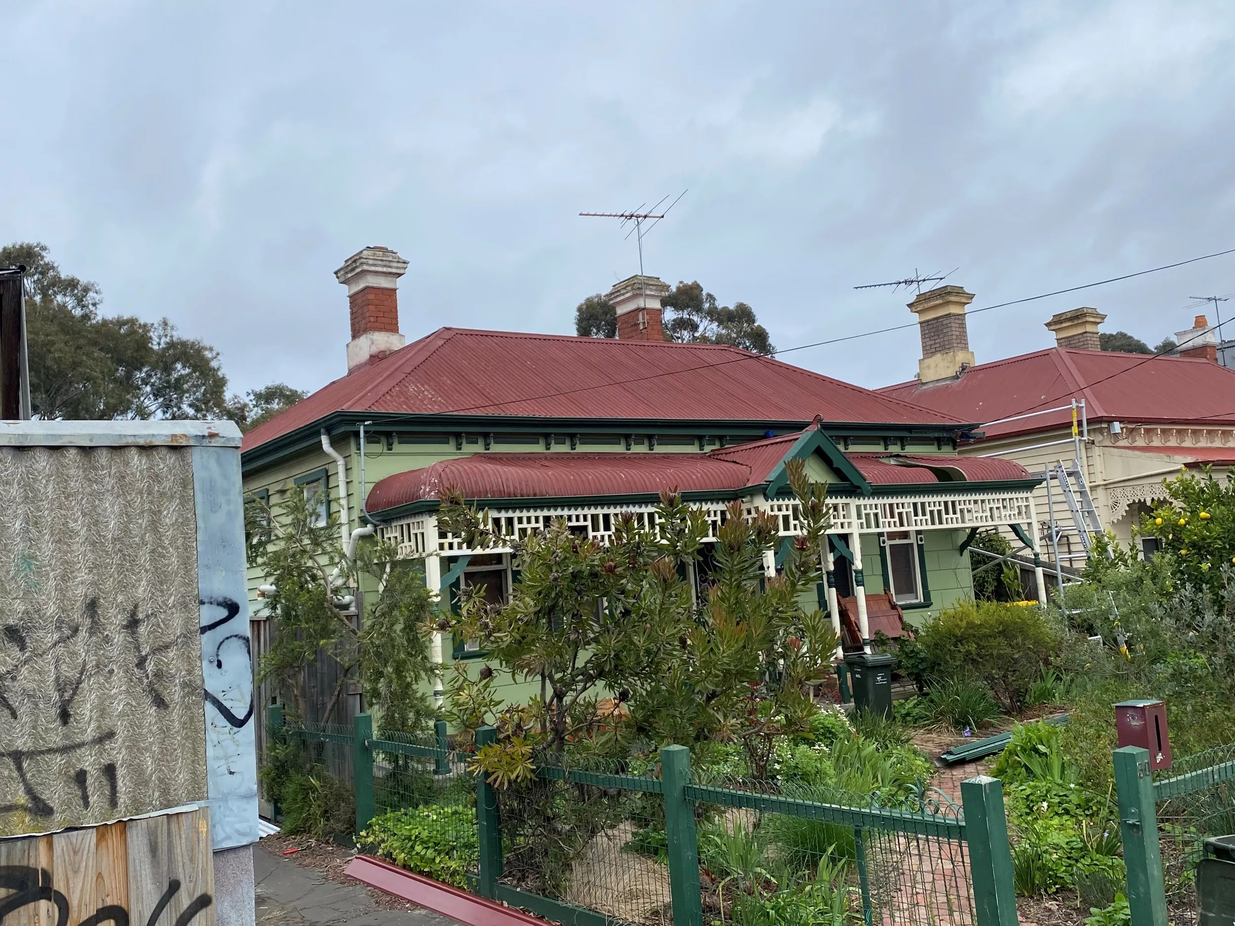 A house with a red tin roof and a chimney, surrounded by lush greenery, before the roof restoration